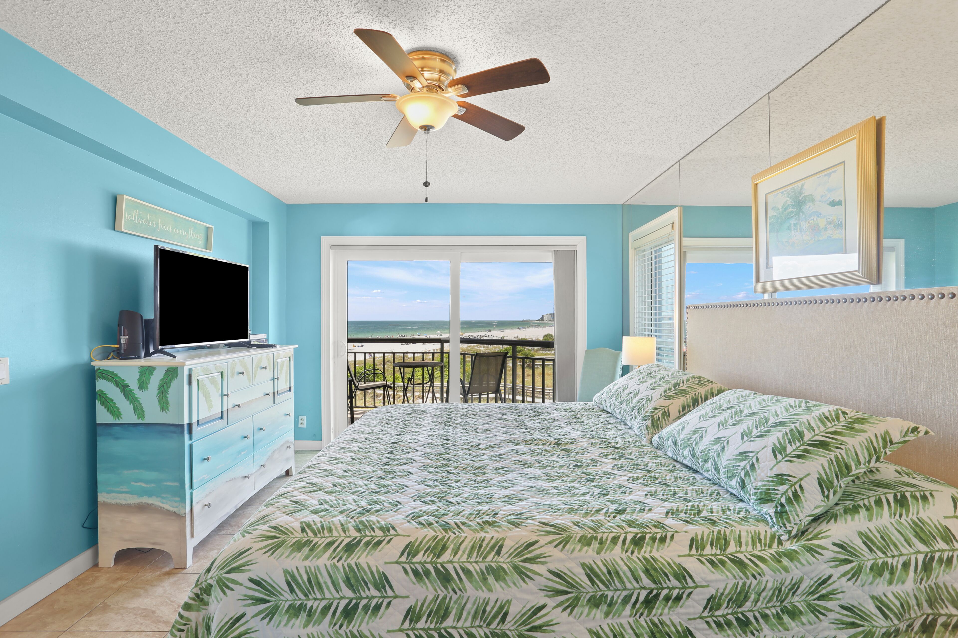 Un dormitorio con cama, ventilador de techo, cómoda, televisor y balcón con vistas.