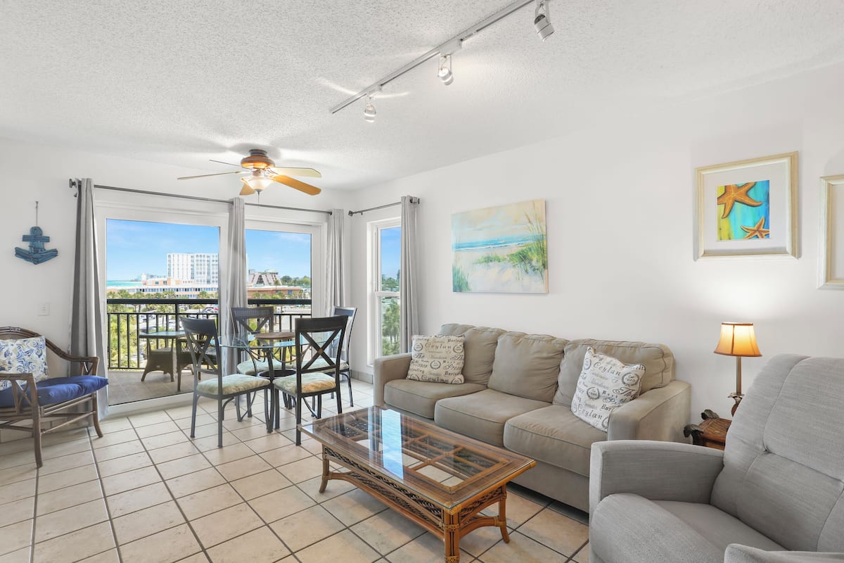 A living room with a glass coffee table, a sofa, a dining table with chairs, a ceiling fan, and a balcony with a view of buildings.