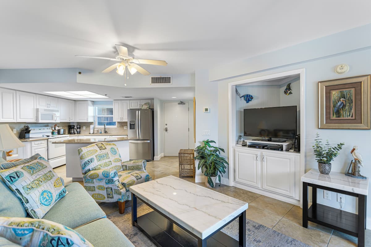 A modern living room with a sofa, a coffee table, and a TV. There is a kitchen area with cabinets and appliances.