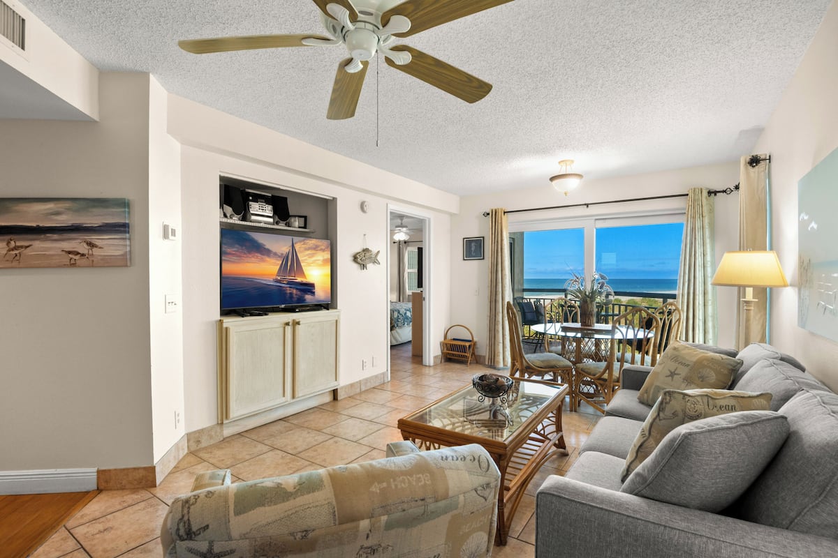 A living room with a television, a ceiling fan, a dining area with chairs, and a view through the window.