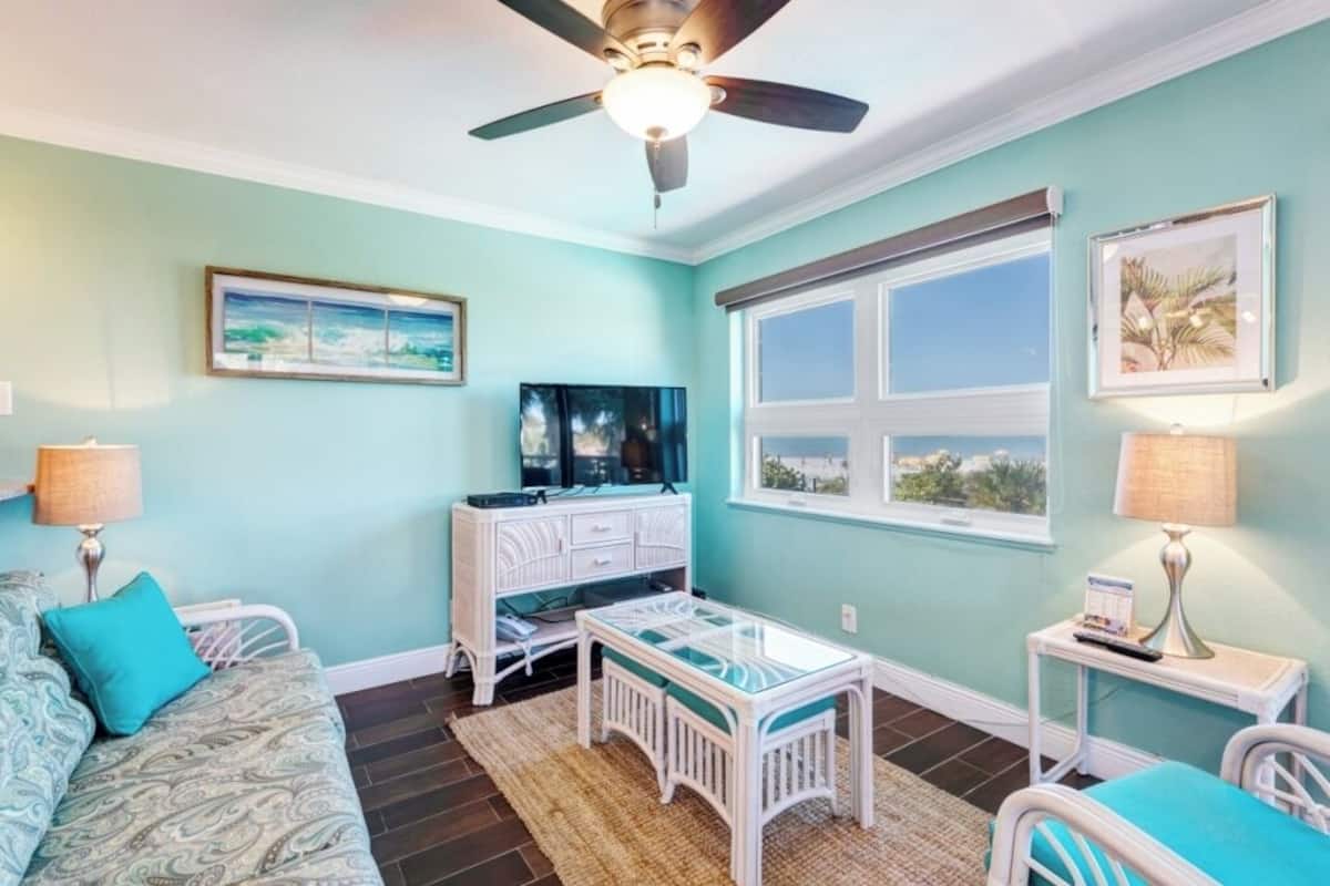 A room with teal walls, a ceiling fan, a bed, a white dresser, a TV, and a window with a view.