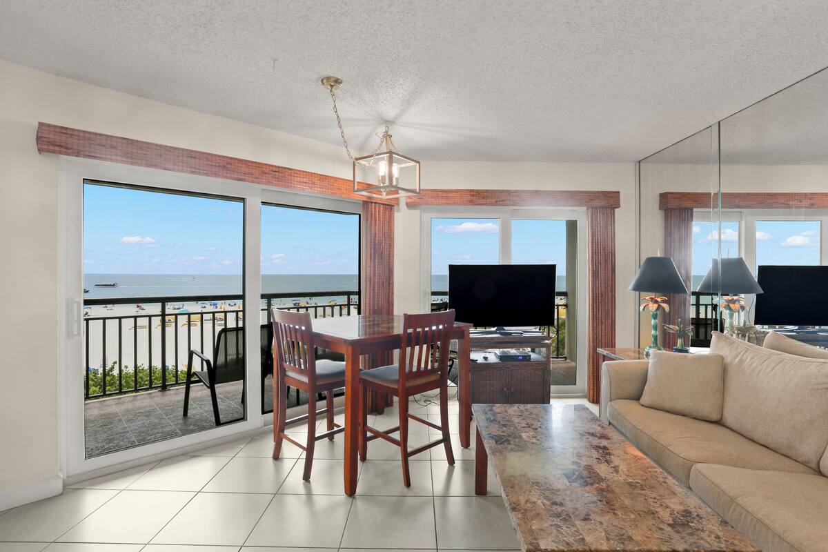 A living room with a large TV, wooden furniture, and a balcony with a view of the sea.