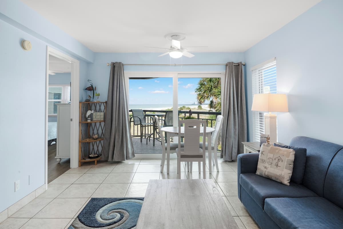 A living room with a blue sofa, a coffee table, a ceiling fan, and a view of the ocean through a glass door.