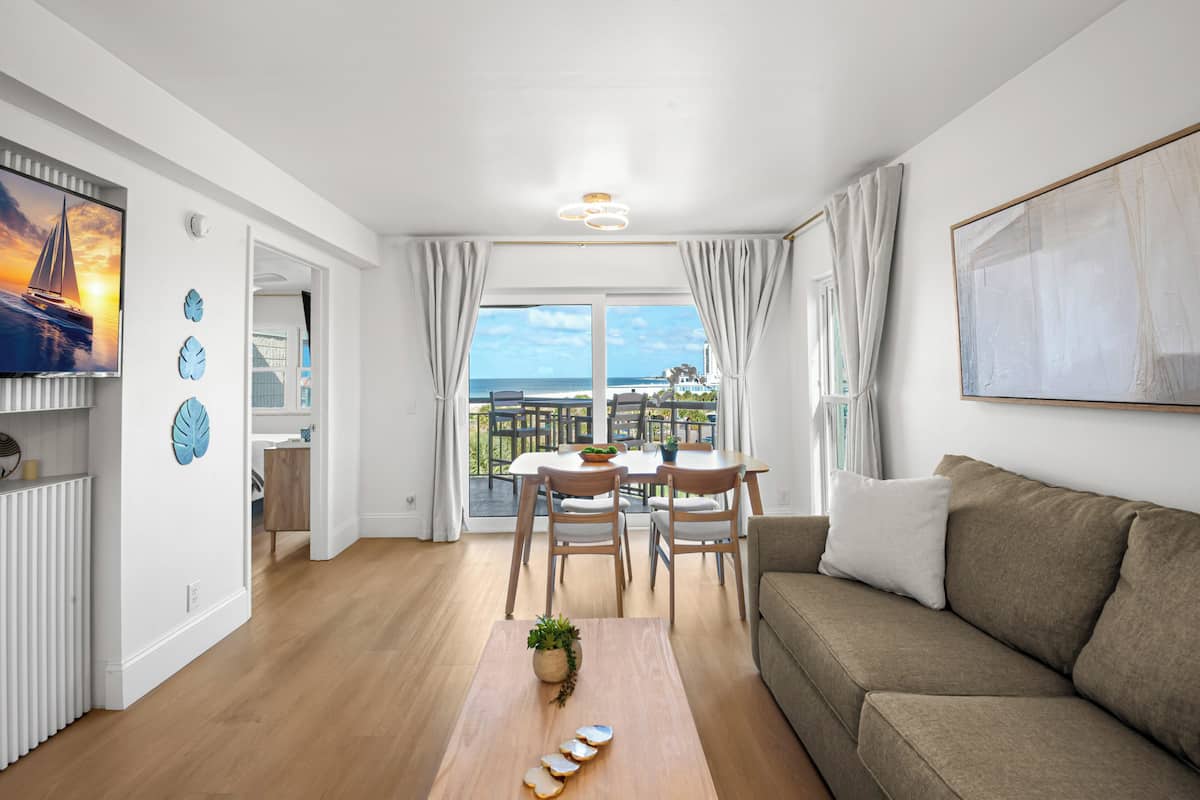A modern living room with a wooden floor, a sofa, a dining table with chairs, a TV, and a balcony with a view.