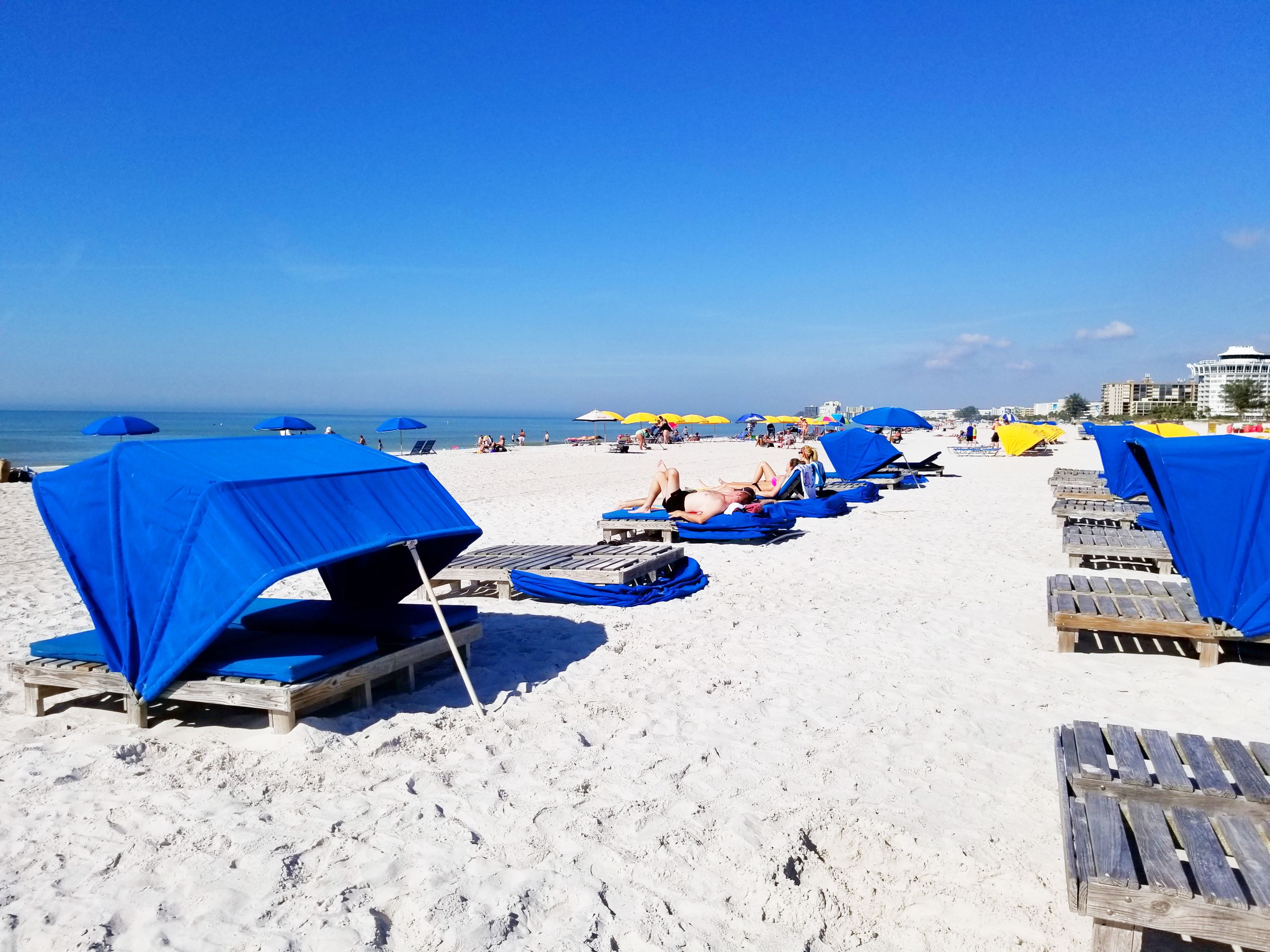 En strand med rader av träsolstolar och blå parasoller, och några personer som solar.