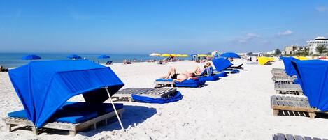 En strand med rader av träsolstolar och blå parasoller, och några personer som solar.