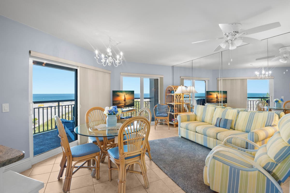 A living room with a sofa, chairs, a dining table, and a TV. There is a balcony with a view of the ocean.