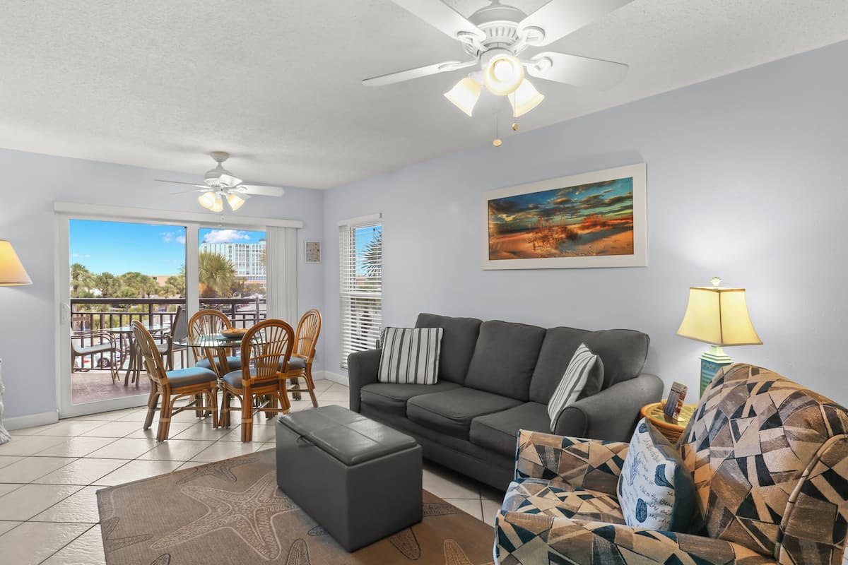 A living room with a sofa, ottoman, and patterned armchair, a ceiling fan, and a dining area with chairs and a table.