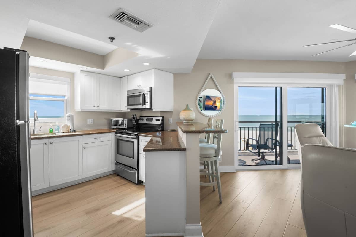 A kitchen with white cabinets, a microwave, and a stove. There is a dining area with a table and chairs, and a balcony with a view of the ocean.