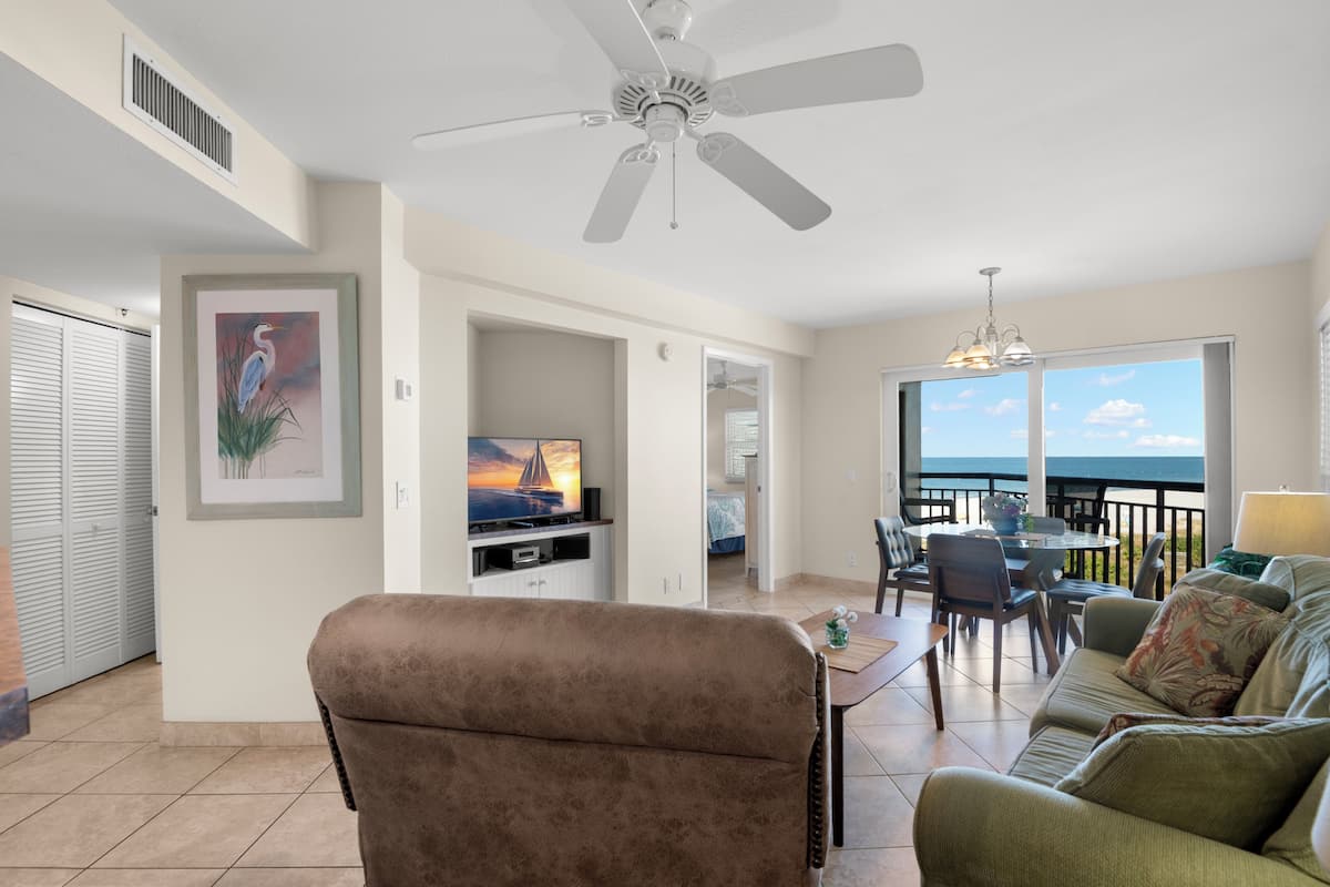 A living room with a TV, a ceiling fan, a dining area, and a balcony with a view.