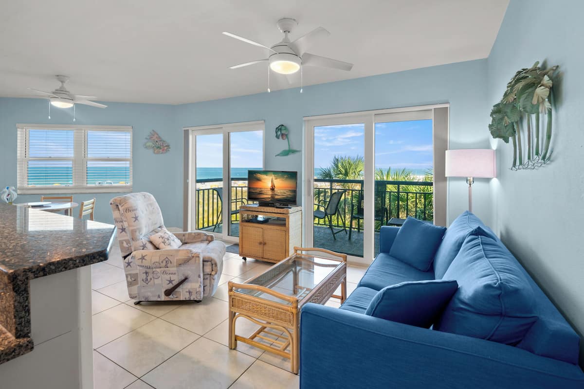 A living room with a blue sofa, a wicker chair, a glass coffee table, a TV, and a balcony with a view of the ocean.