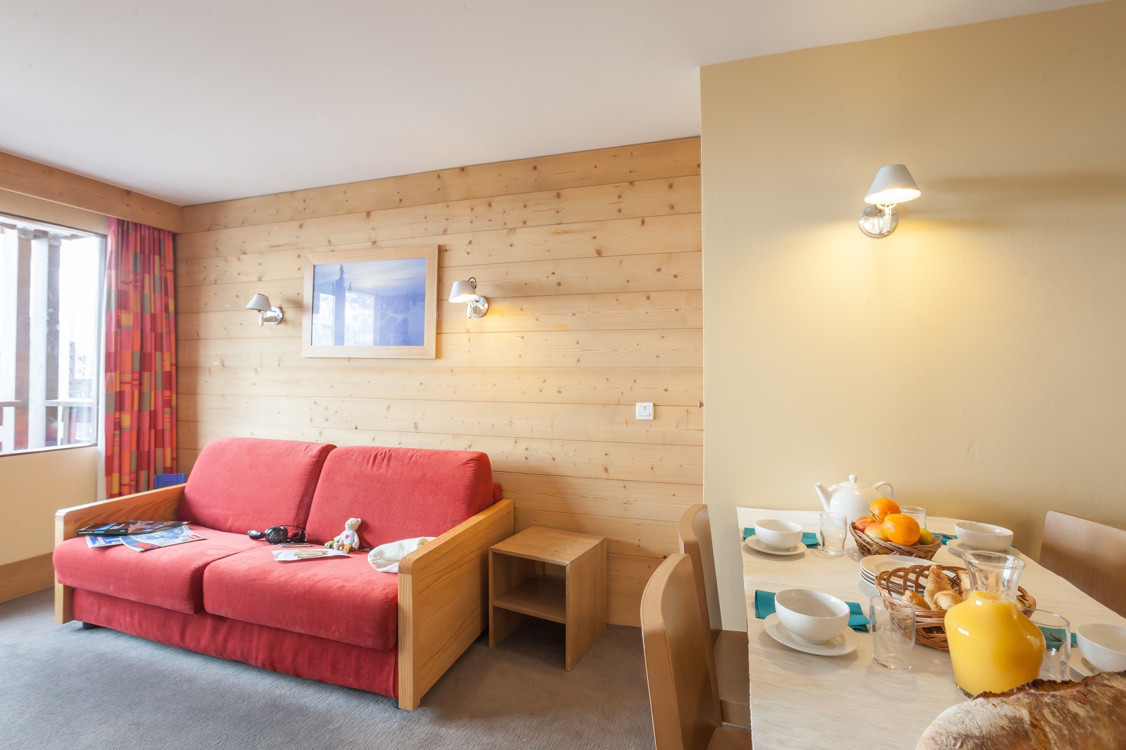 A cozy room with a wooden wall, a red sofa, a small table, and a breakfast setup with cups, a jug, and a basket of fruit.