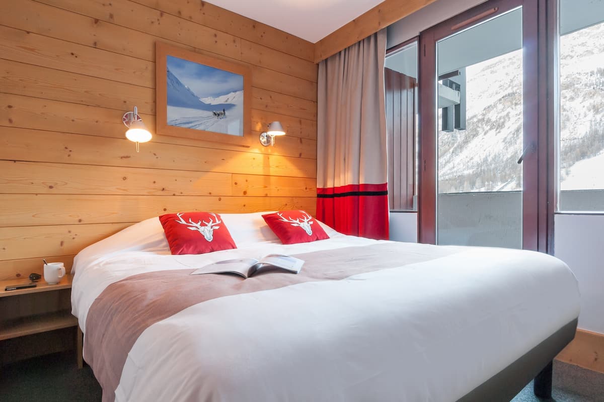 Ein Zimmer mit Holzwänden, einem Bett, zwei Wandleuchten, einem mit Vorhängen versehenen Fenster und einem gerahmten Bild.