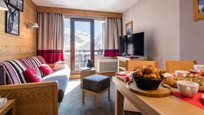 Una habitación acogedora con paredes de madera, un sofá, un televisor, una mesa de comedor con pasteles y vistas a montañas nevadas.