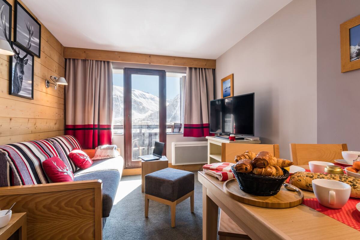 Ein gemütliches Zimmer mit Holzvertäfelung, einer Couch, einem Fernseher, einem Esstisch mit Gebäck und Blick auf schneebedeckte Berge.