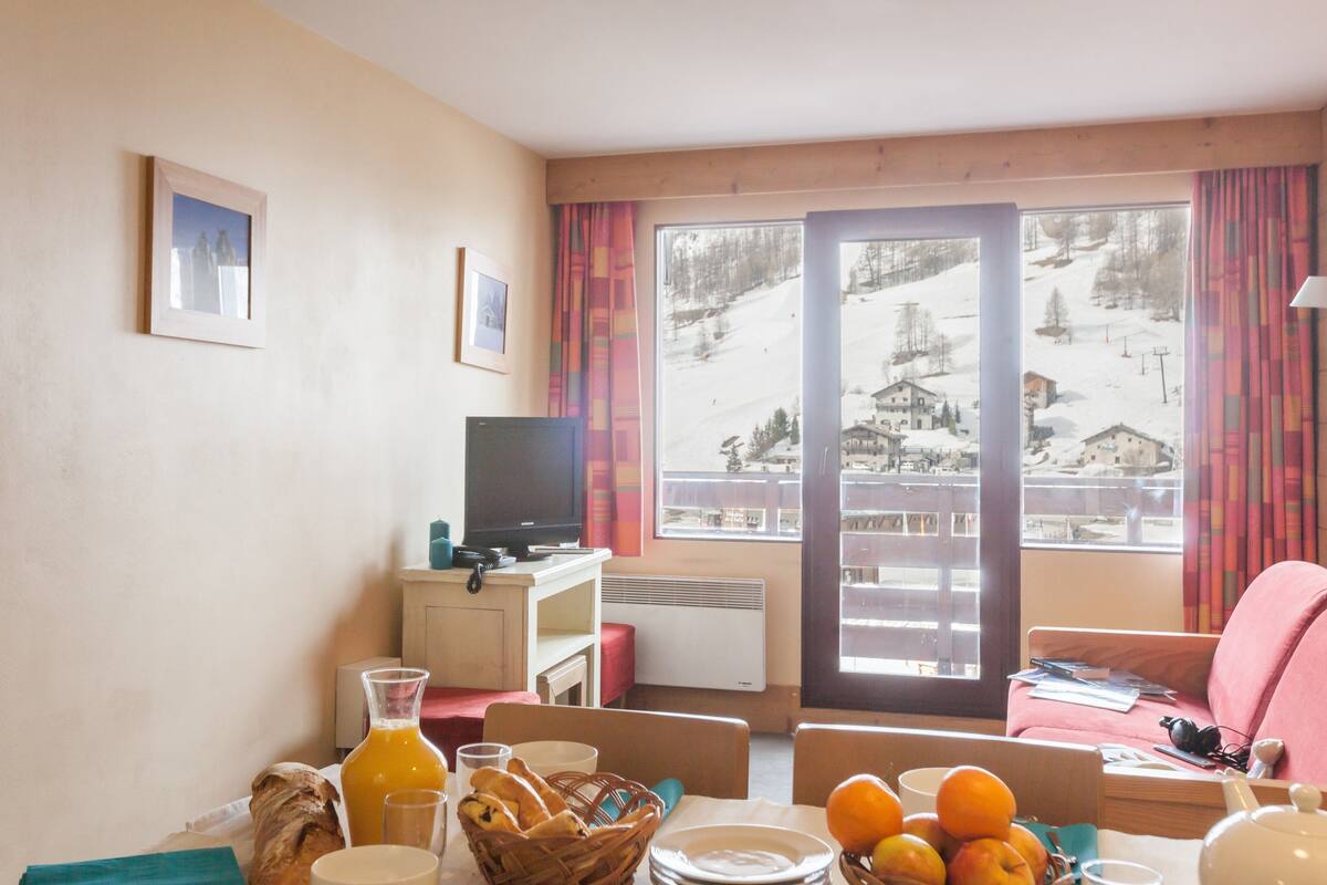 Ein gemütliches Zimmer mit Esstisch, gedeckten Früchten und einer Orangensaftflasche, einem Fernseher und Blick auf eine verschneite Berglandschaft.