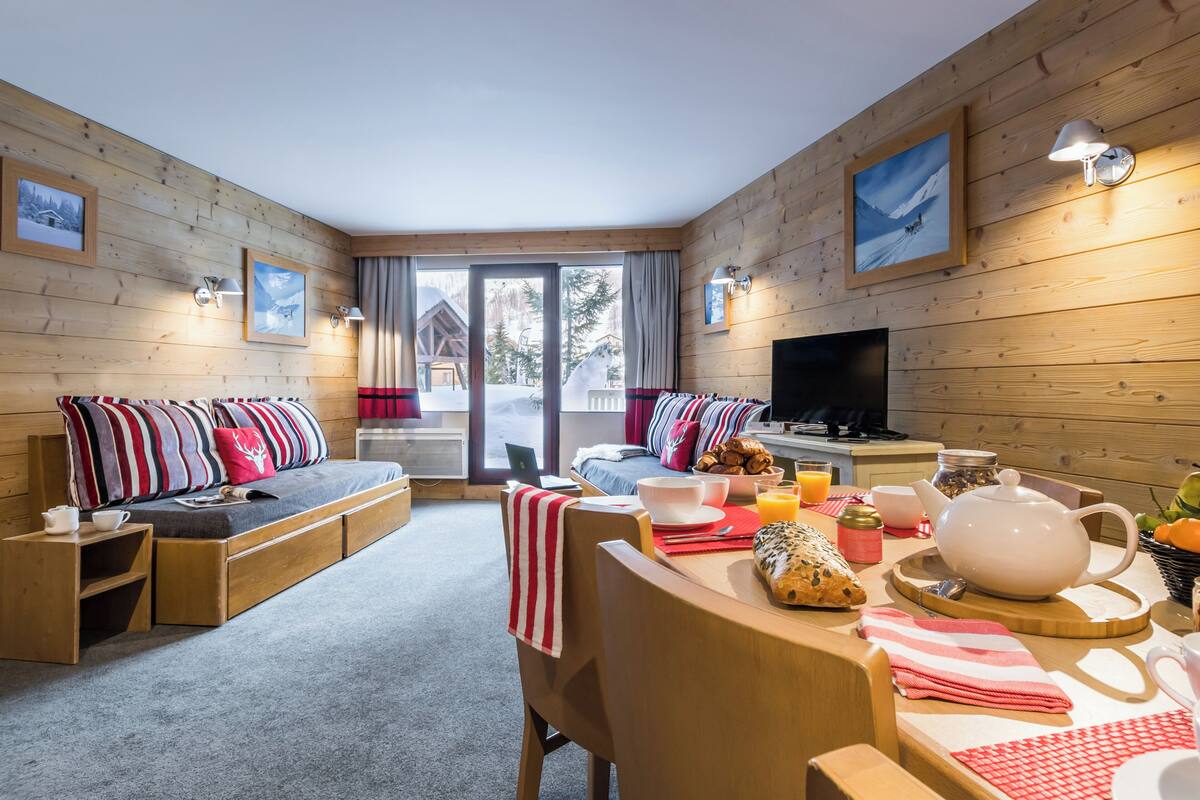 Ein gemütliches Zimmer mit einer Couch, einem Esstisch und einem Fernseher. Holzverkleidungen und ein großes Fenster sind vorhanden.