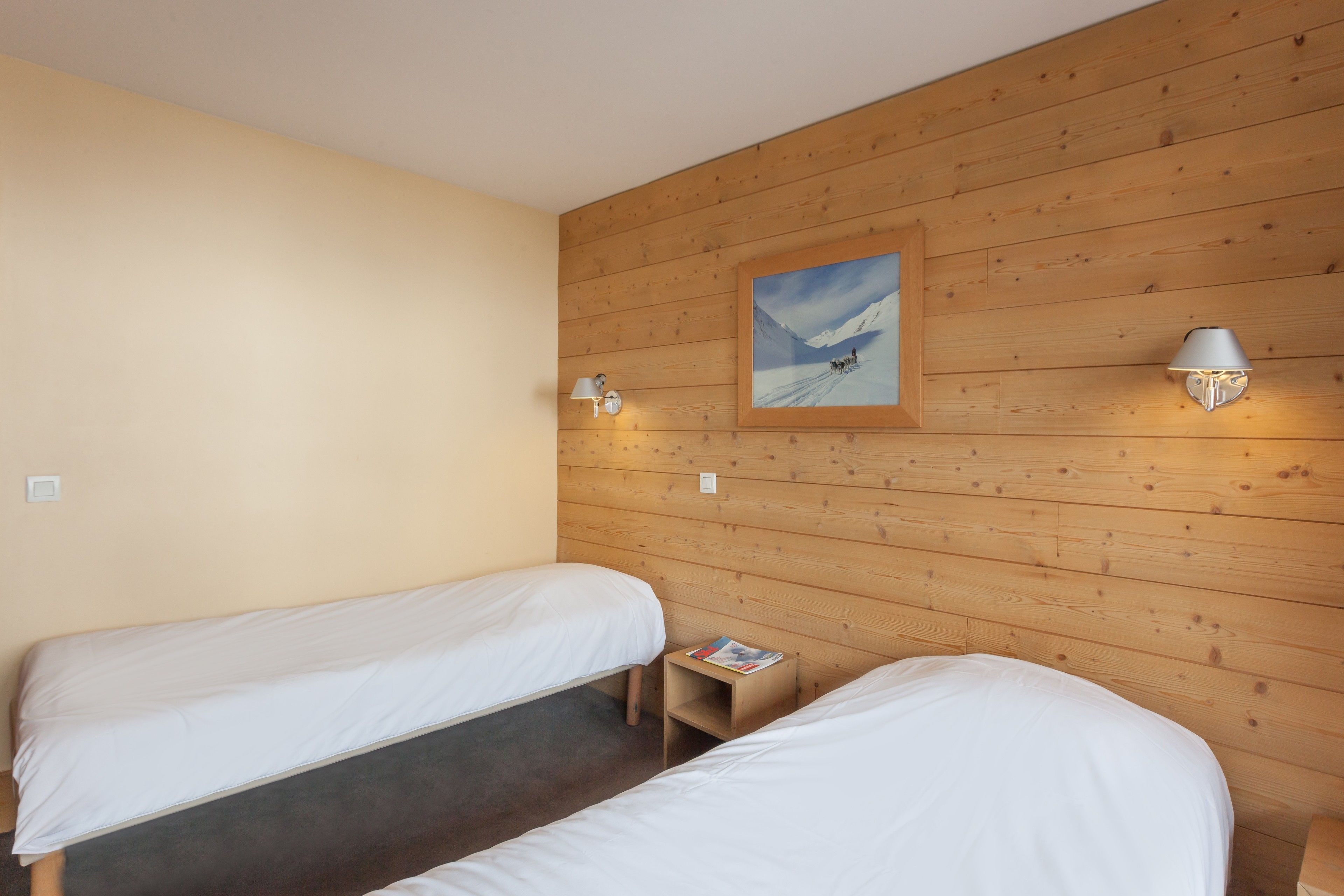 A room with two single beds, wooden panel walls, a painting, and two wall-mounted lamps.