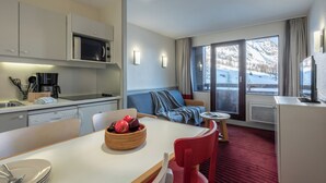A modern hotel room with a kitchen, dining area, and a large window with a mountain view. - Résidence Pierre & Vacances La Daille (Val-d'Isere)