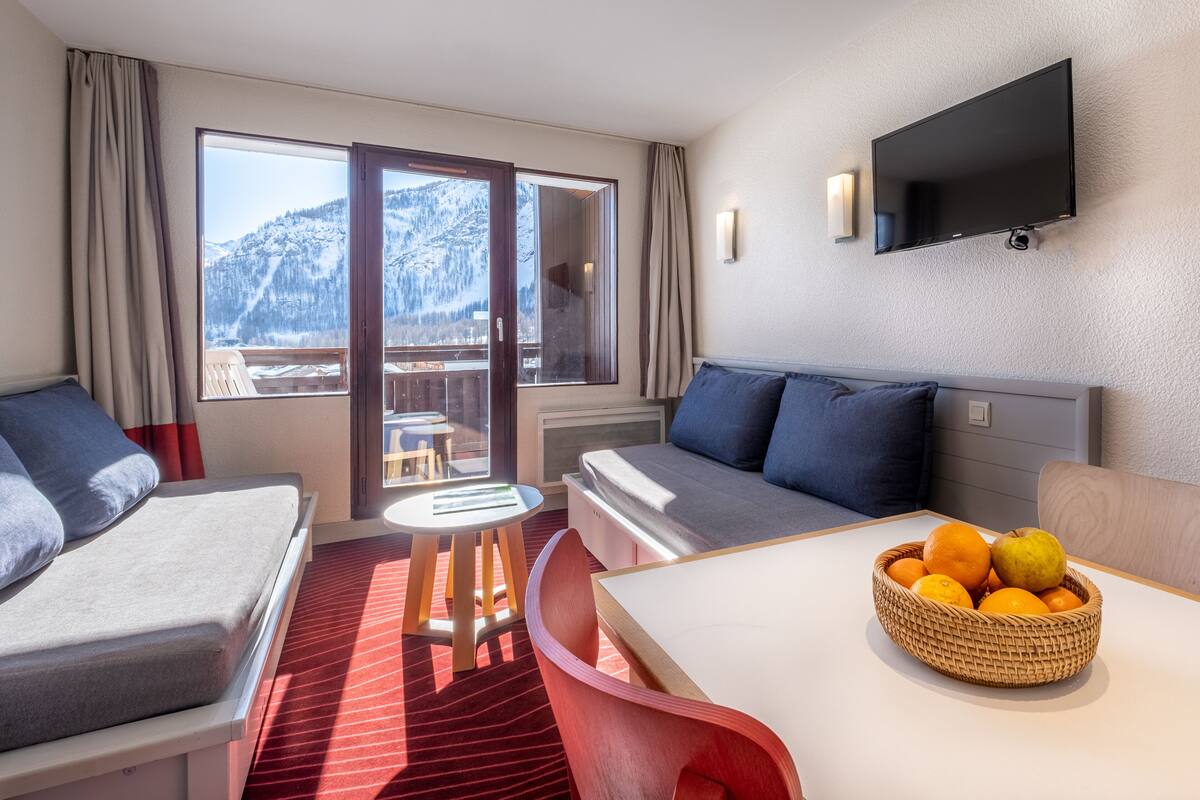 Une chambre d’hôtel avec un lit, un canapé, une télévision, une table avec des fruits et un balcon offrant une vue sur la montagne.