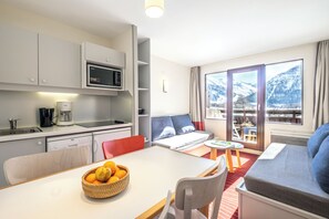 A modern hotel room with a kitchen, dining area, and a sofa with a mountain view. - Résidence Pierre & Vacances La Daille (Val-d'Isere)
