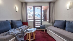 A room with a sofa, a small table, a chair, and a view of mountains through a door. - Résidence Pierre & Vacances La Daille (Val-d'Isere)
