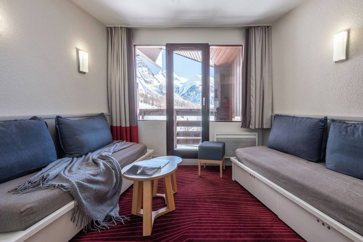 Une pièce comprenant un canapé, une petite table, une chaise et une vue sur les montagnes, visible par la porte.