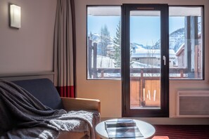 Una habitación con sofá, una mesa, una ventana con vistas a un paisaje nevado, una cortina y una lámpara fijada a la pared.