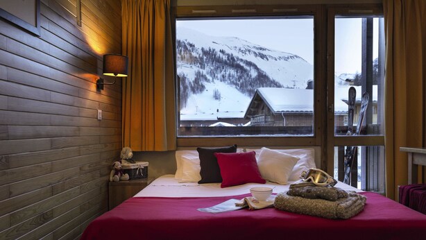 A hotel room with a wooden wall, a large window with a snowy mountain view, a bed with a red blanket, pillows, a teddy bear, and a ski helmet on the bed.