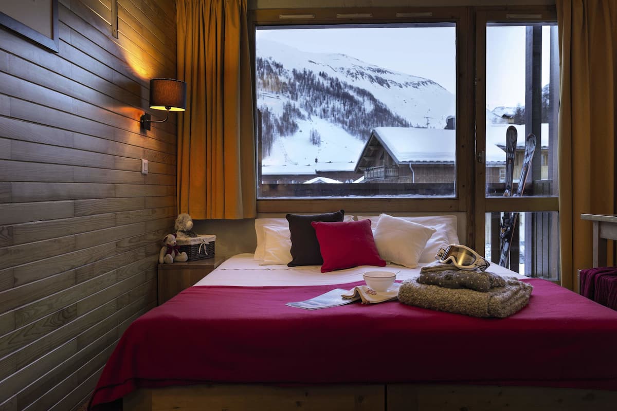 Ein Hotelzimmer mit Holzwand, einem großen Fenster mit Blick auf einen verschneiten Berg, einem Bett mit roter Decke, Kissen, einem Teddybären und einem Skihelm auf dem Bett.