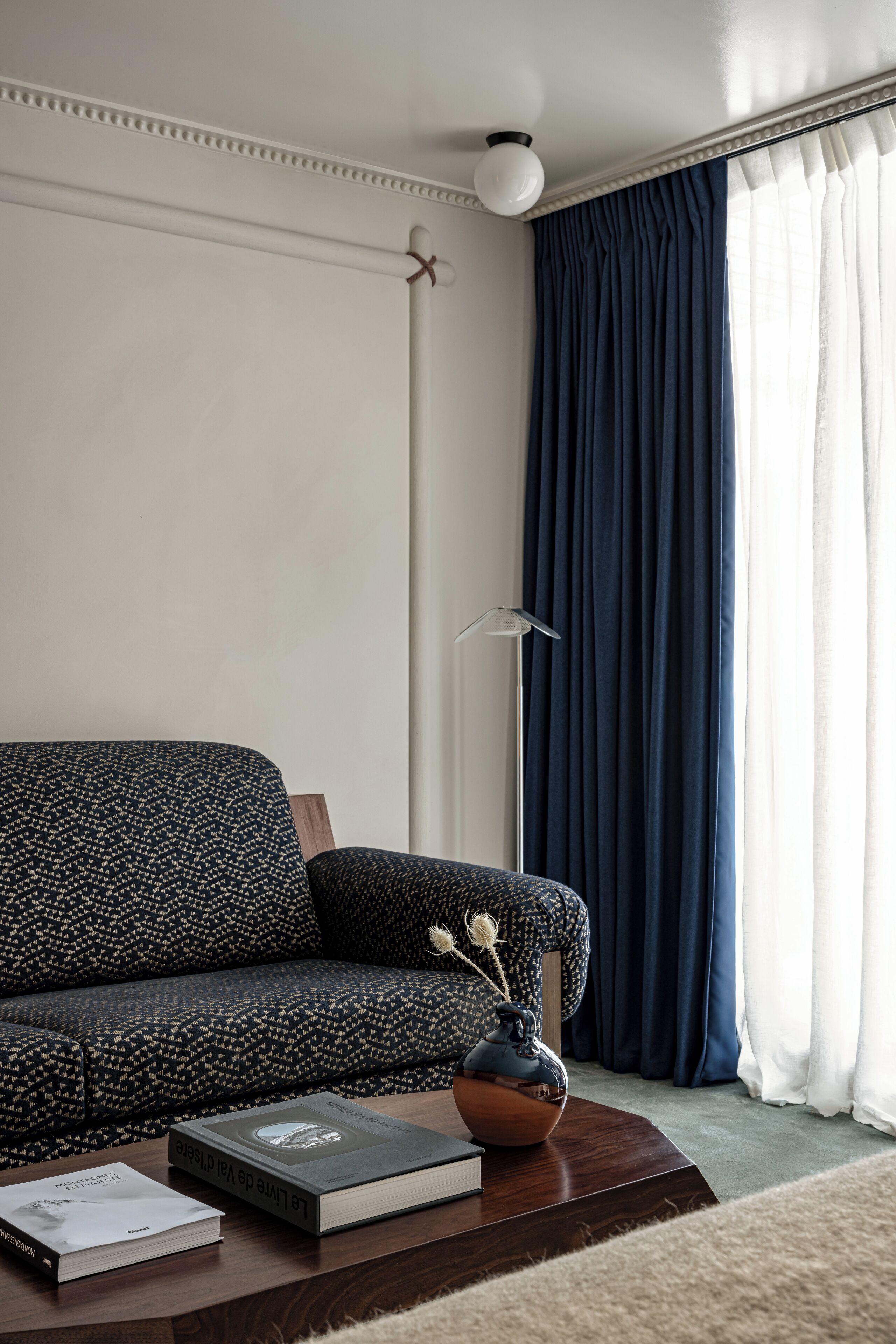 En stue med en mørk sofa med et mønster, et træsofabord med en bog og en vase, samt en gulvlampe.