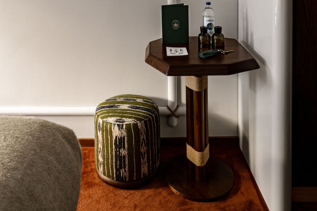 A corner of a room with a round, striped ottoman, a small wooden table with a green box and bottles, and a white wall.