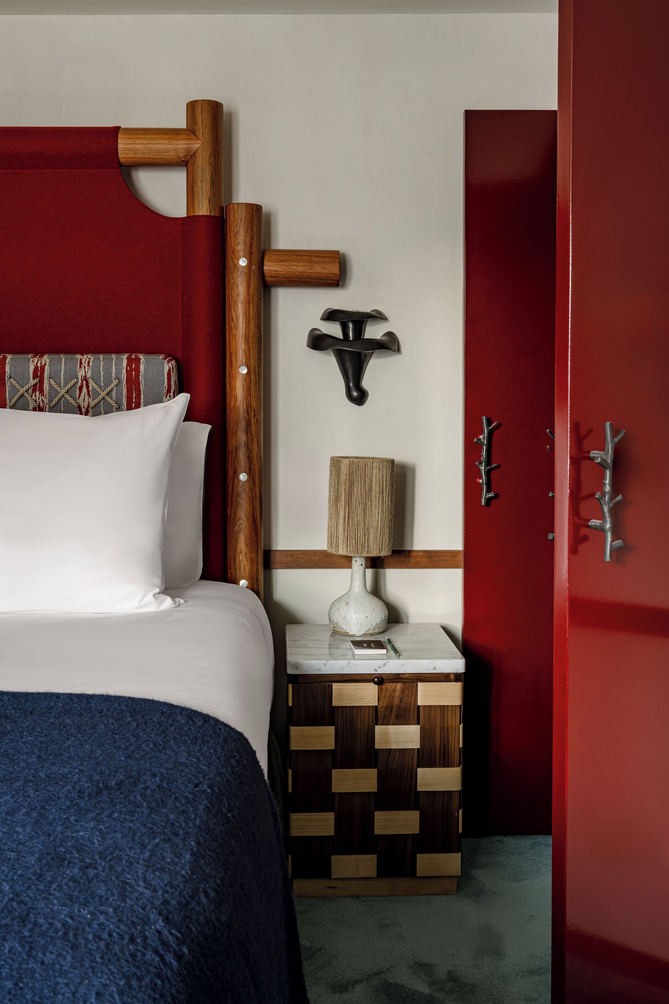 A neatly arranged hotel room with a bed, a bedside table with a lamp, a wall-mounted light fixture, and a red wardrobe.