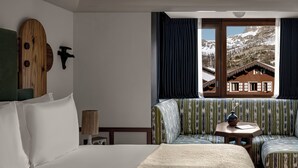 Habitación de hotel con una cama grande, un sofá verde a rayas, una mesita redonda y una ventana con vistas a una montaña y un edificio.