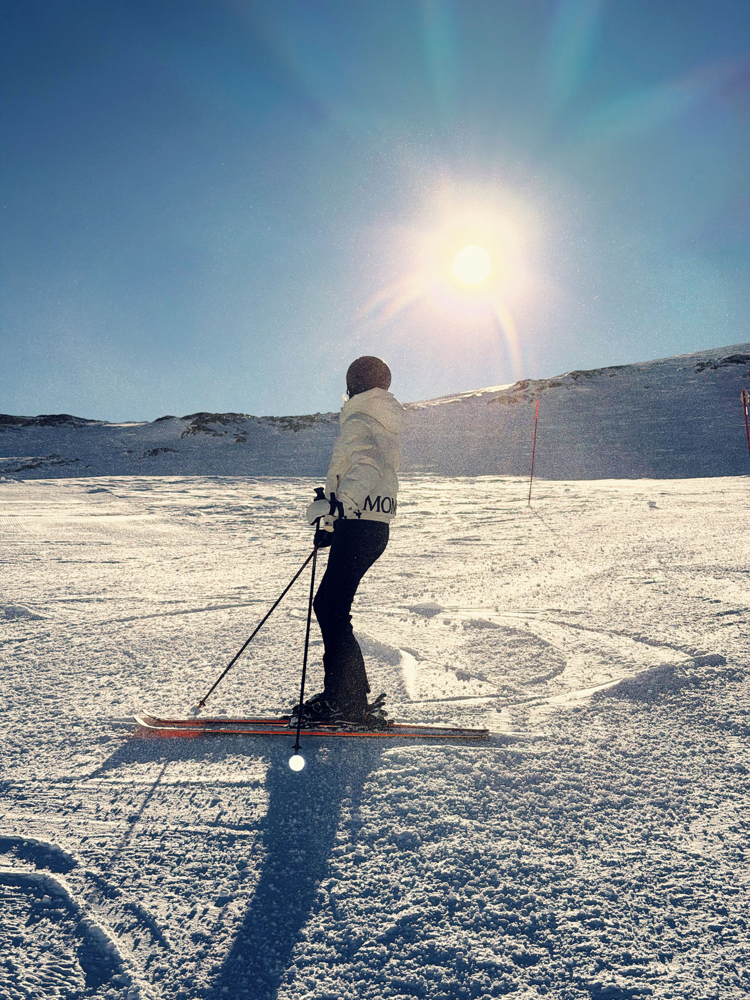Une personne fait du ski sur une pente enneigée, avec un ciel dégagé et le soleil en arrière-plan.