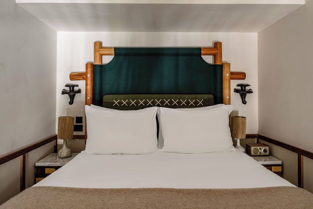 Uma cama bem arrumada com cabeceira de madeira, duas mesas de cabeceira com luminárias e um rádio.