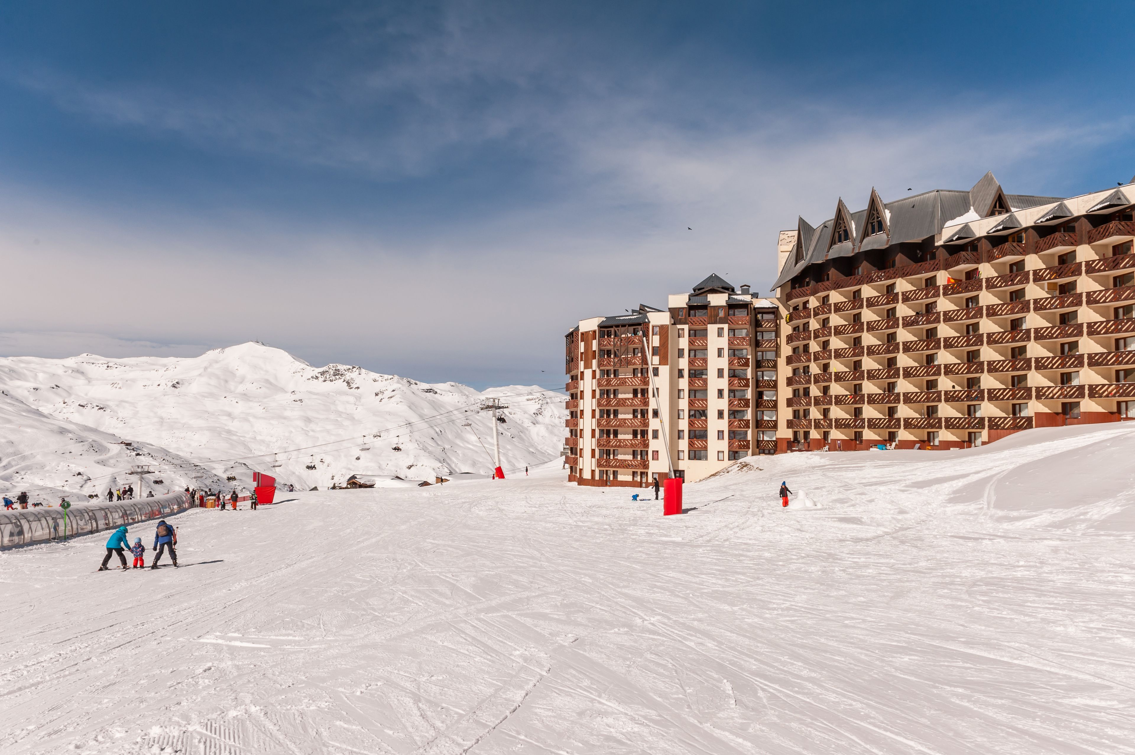A snow-covered ski resort with a large hotel featuring multiple balconies and a ski lift in the background.