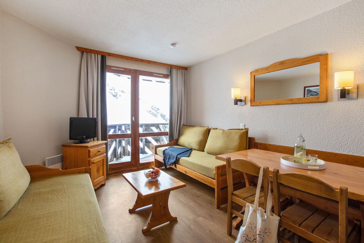 A cozy room with a sofa, a wooden table, a TV, and a balcony with a view of mountains.