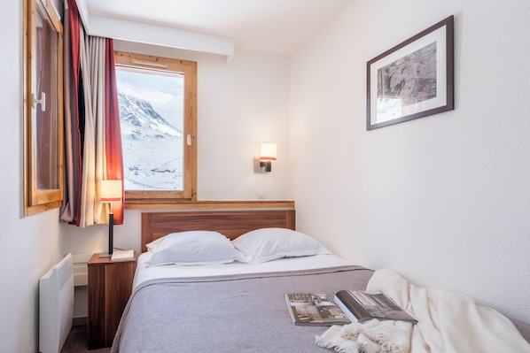 Un dormitorio con una cama, una mesita de noche, una lámpara, un cuadro enmarcado y una ventana con cortinas.