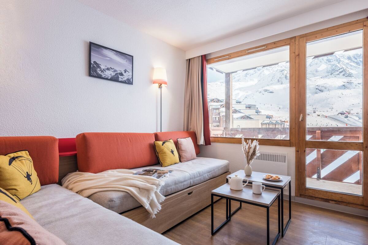 A modern hotel room with a sofa, a small table with a teapot and cups, a lamp, and a window with a mountain view.
