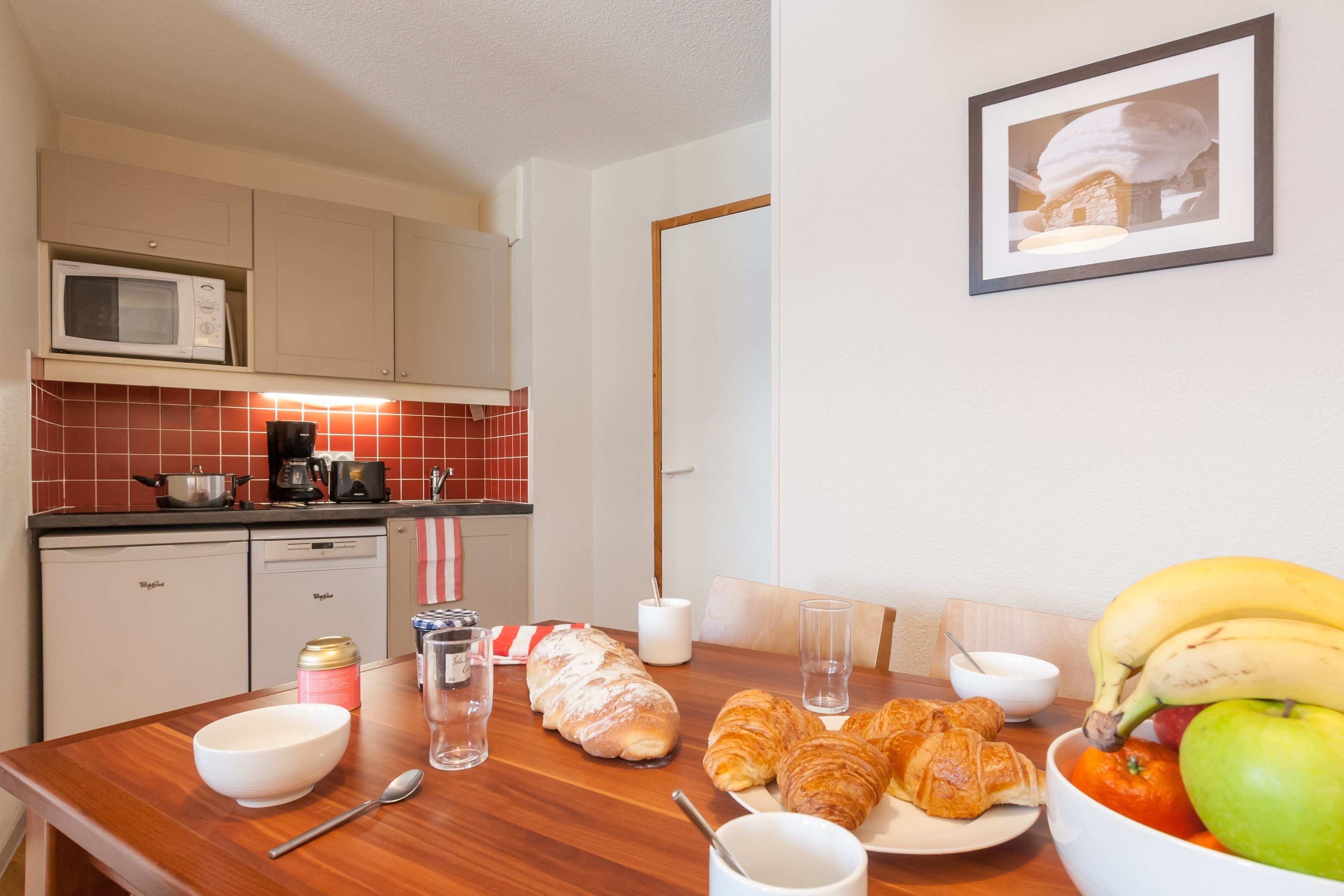A kitchen with a microwave, toaster, and coffee maker. There is a dining table with croissants, a bowl of fruit, and a framed picture on the wall.