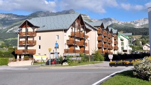 Un edificio de varios pisos con balcones, un cartel con los números de las habitaciones y montañas al fondo.