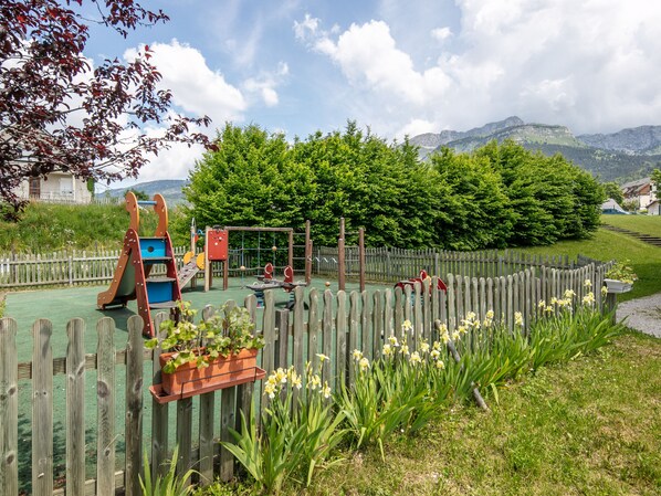 A playground with various play structures, surrounded by a wooden fence and lush greenery.