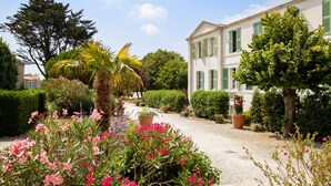 A garden with a gravel path leading to a white building with green shutters. - Résidence Le Palais des Gouverneurs Pierre & Vacances (Saint-Martin-de-Re)