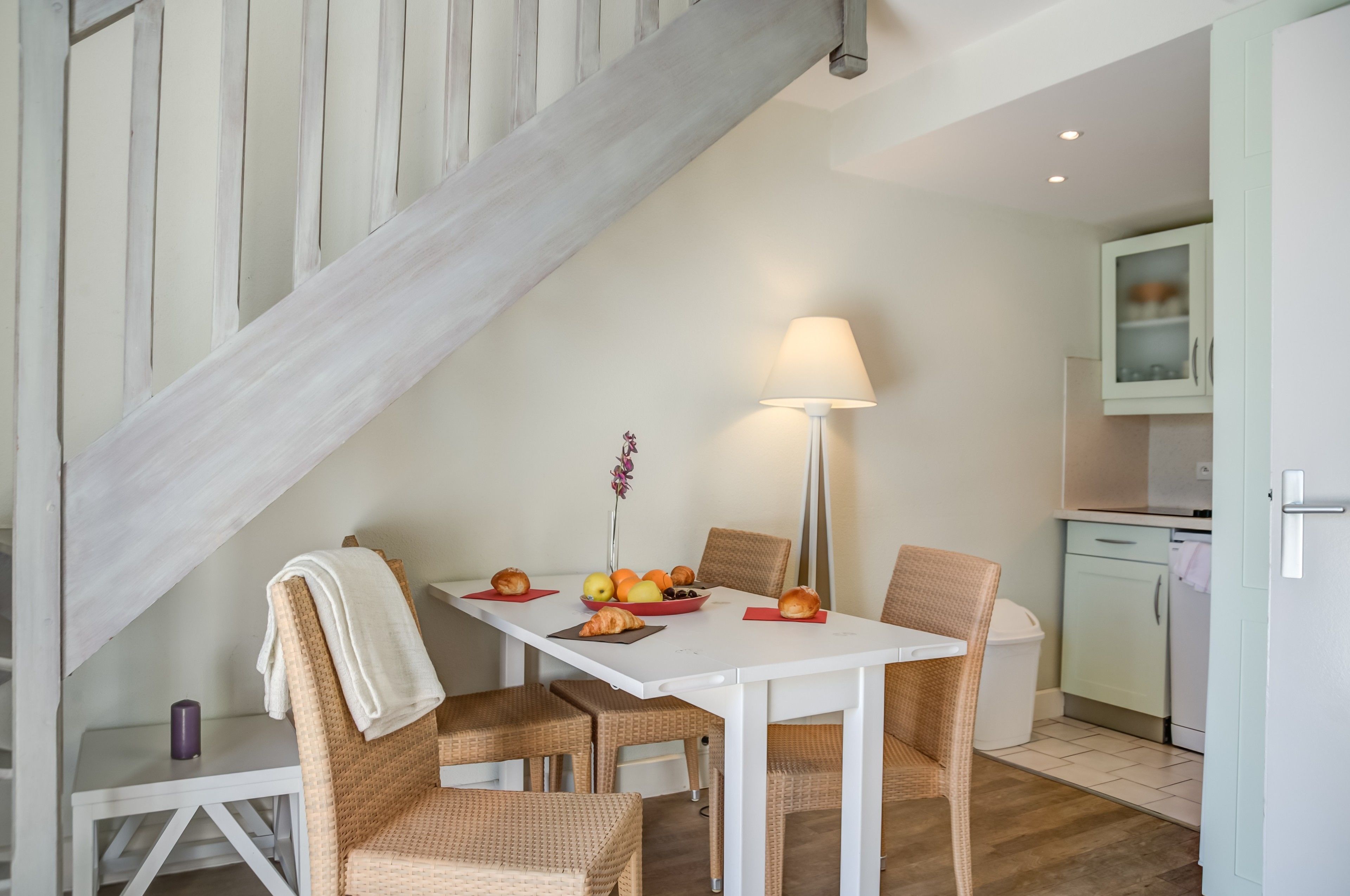 A dining area with a white table, wicker chairs, a lamp, and a fruit bowl.