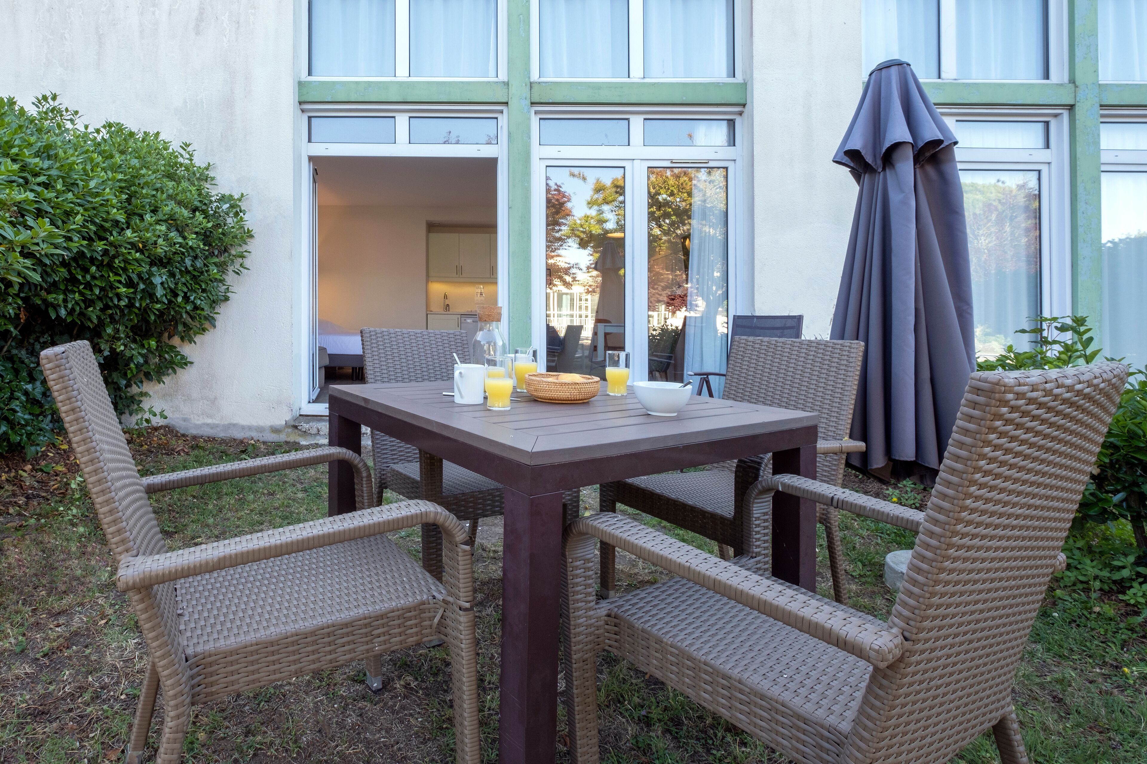 An outdoor dining area with wicker chairs and a table, a grey umbrella, and a view of the house's interior through large windows.