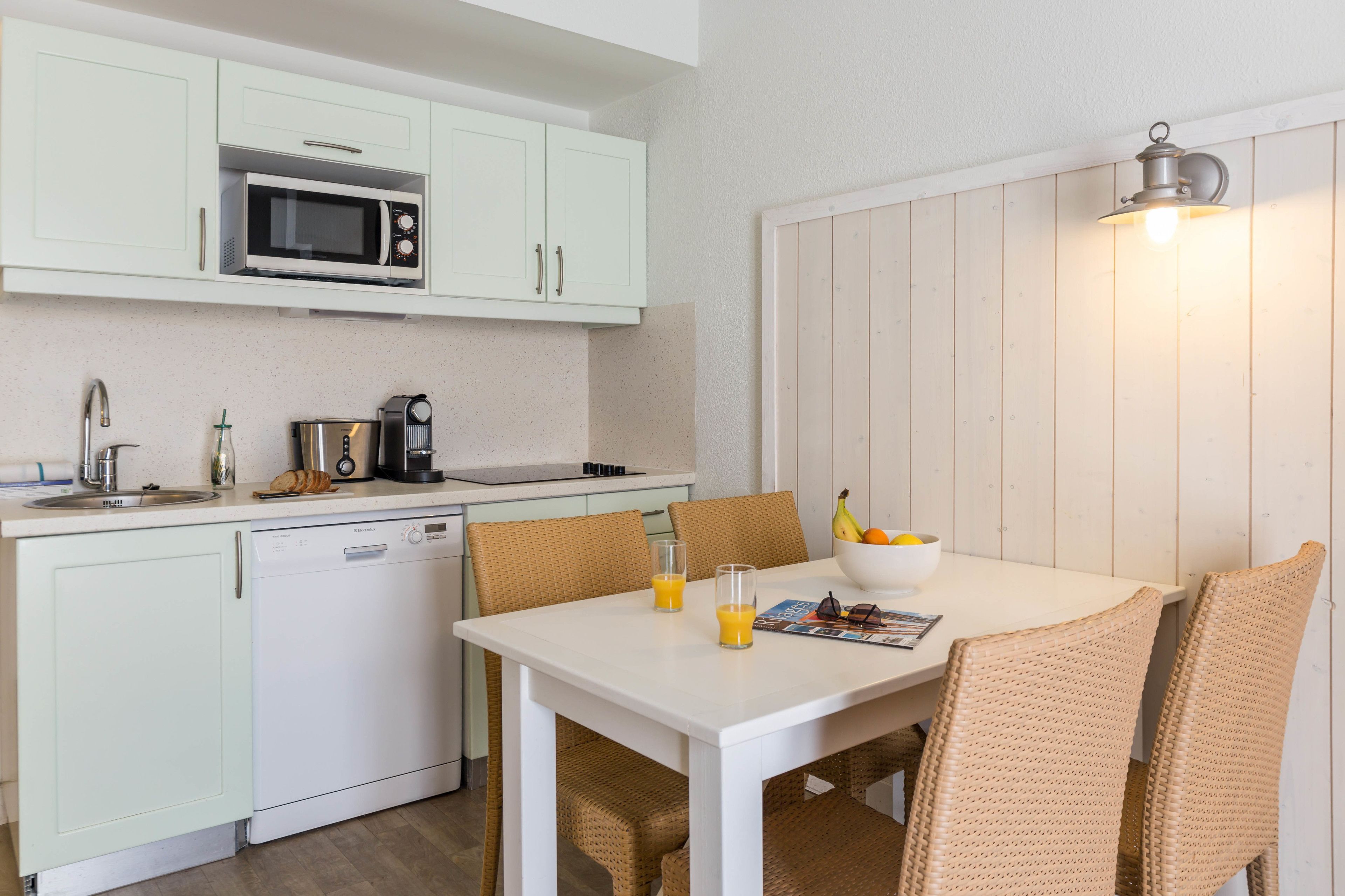 A compact kitchen with white cabinets, a microwave, a coffee maker, and a dishwasher. There is a dining table with wicker chairs and a bowl of fruit.