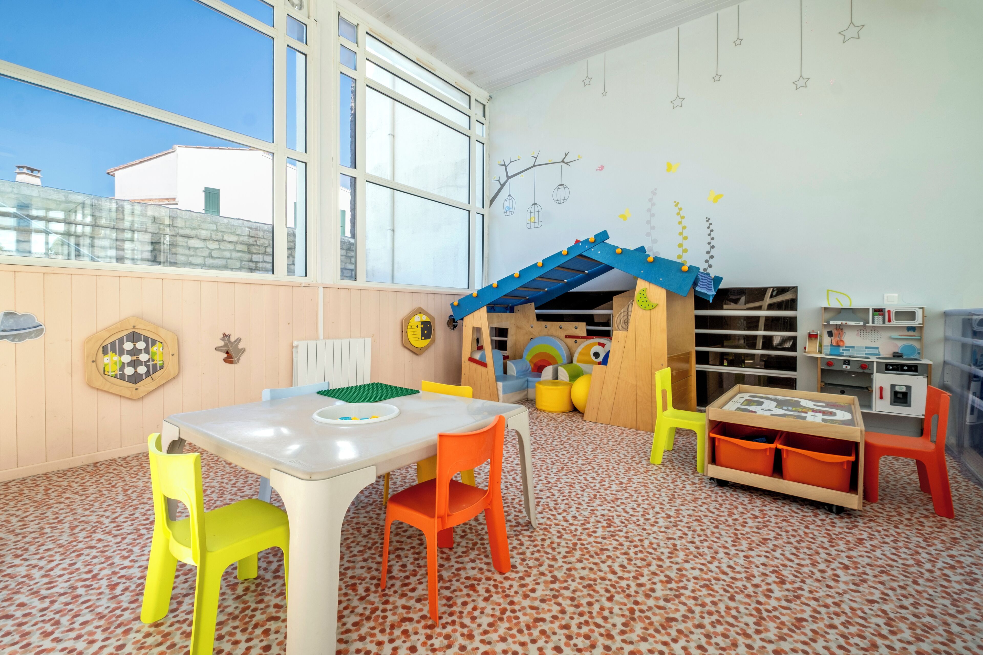 A children's playroom with a large window, wooden play structures, and a white table with chairs.