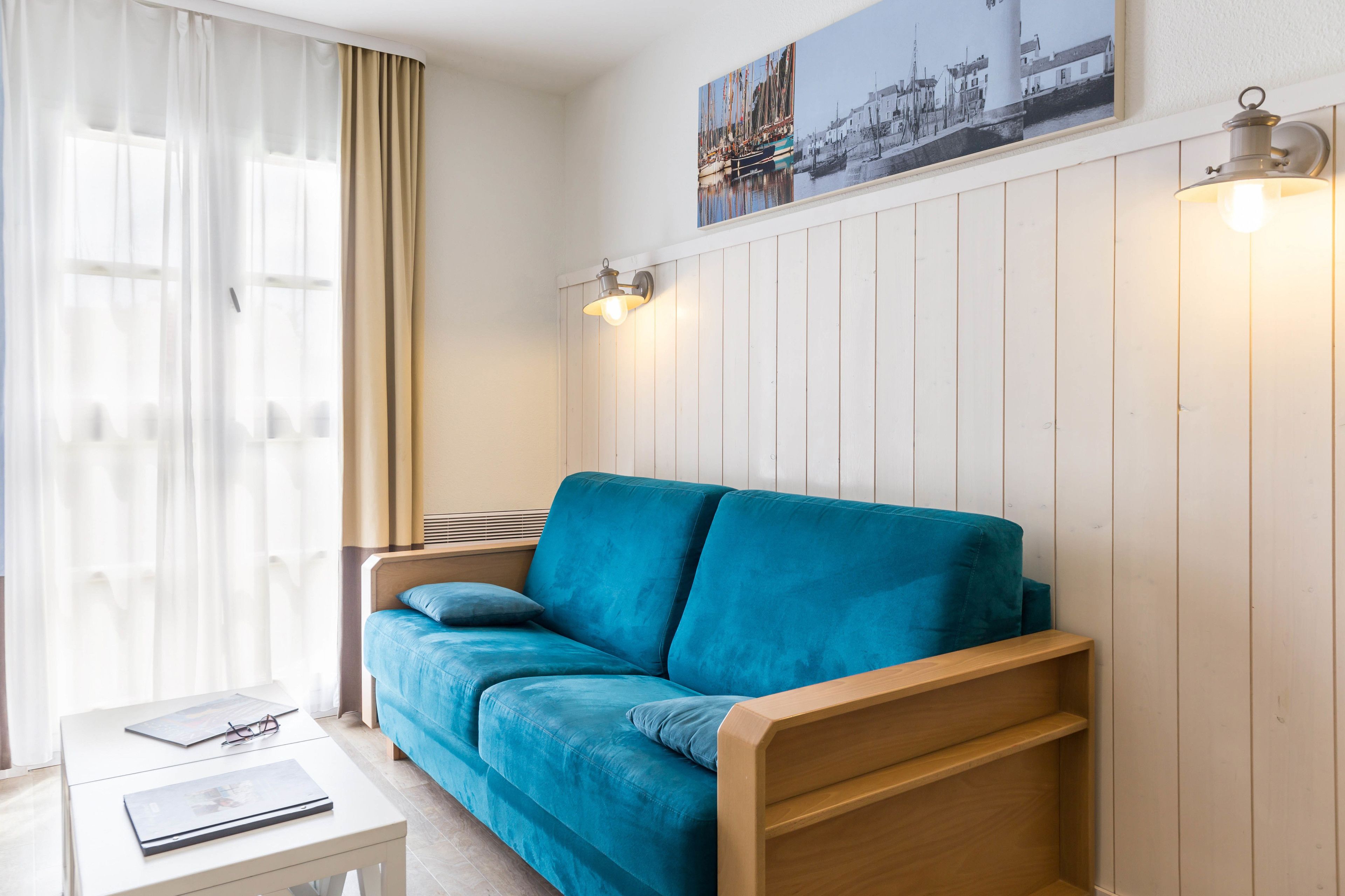 Ein Zimmer mit einem blauen Sofa, einem kleinen weißen Tisch mit Buch und Gläsern, einem Fenster mit durchscheinenden Vorhängen und einem an der Wand hängenden Bild.
