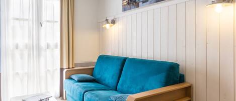 A room with a blue sofa, a small white table with a book and glasses, a window with sheer curtains, and a wall-mounted picture.